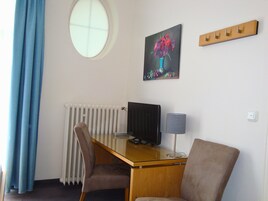 Ein Zimmer mit einem Rundfenster, einem Schreibtisch mit Computer, einem Stuhl, einem Heizkörper und einer Wandleuchte.