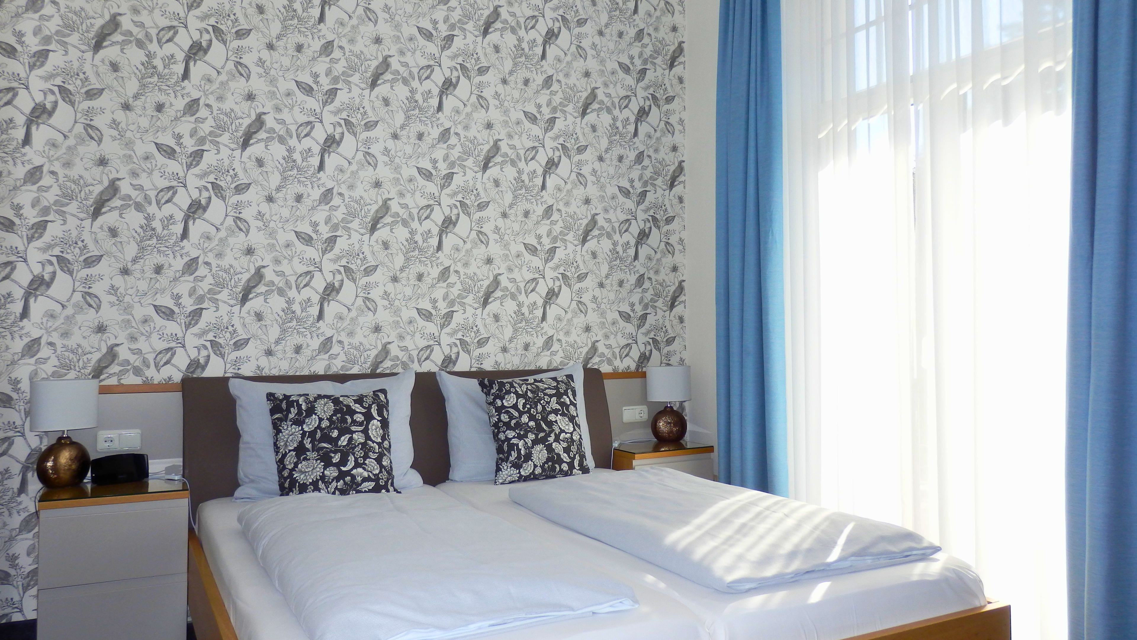Habitación de hotel con papel tapiz floral, una cama con ropa de cama blanca, dos mesitas de noche con lámparas y una ventana con cortinas azules.