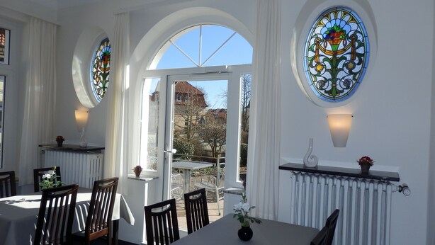 A dining area with a table and chairs, a view through an open door, and two wall-mounted lamps. - Hotel Bellevue (Hamelin)