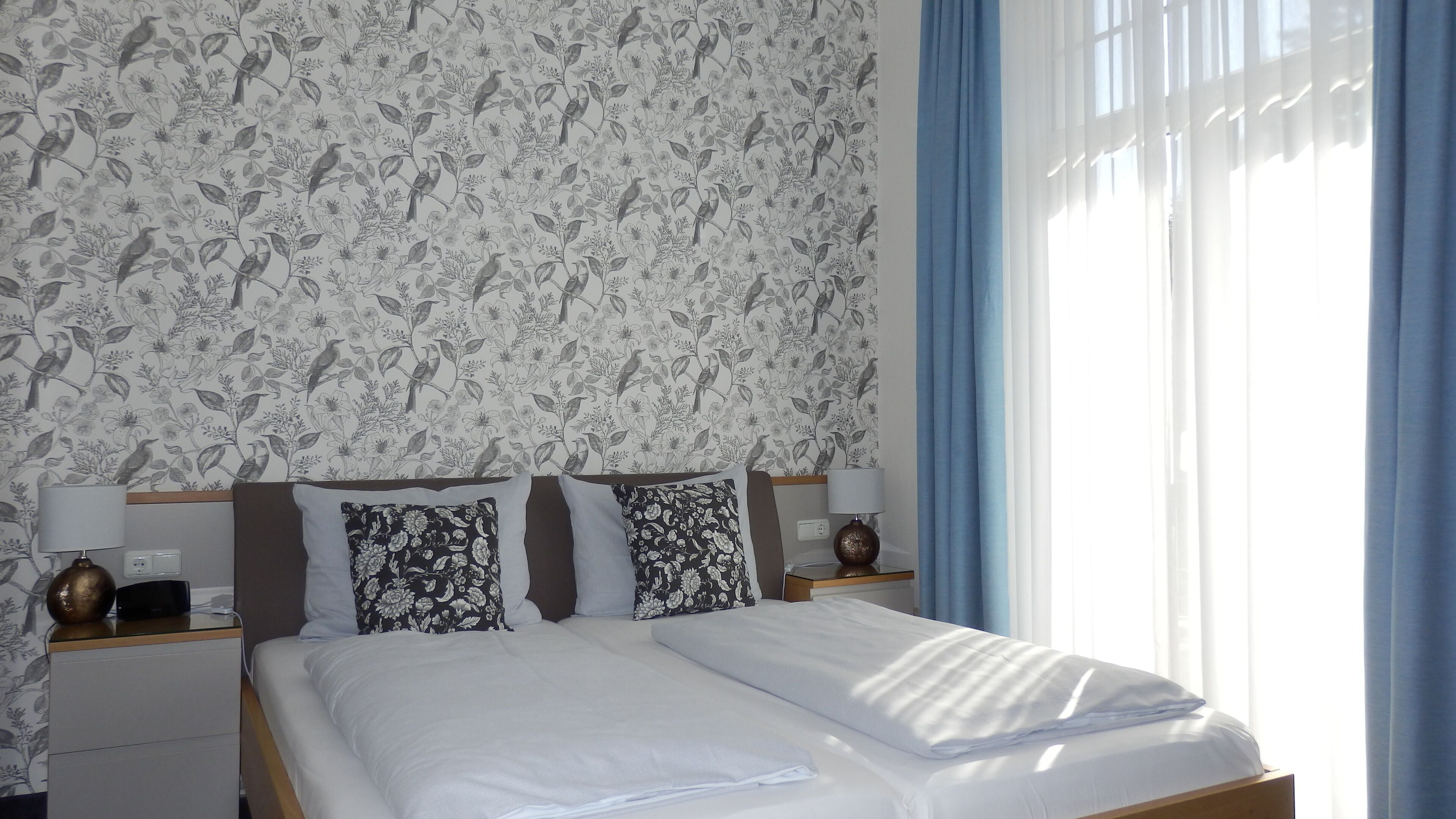 Una cama bien hecha con sábanas blancas, dos mesitas de noche con lámparas, y un estampado floral en el papel tapiz.