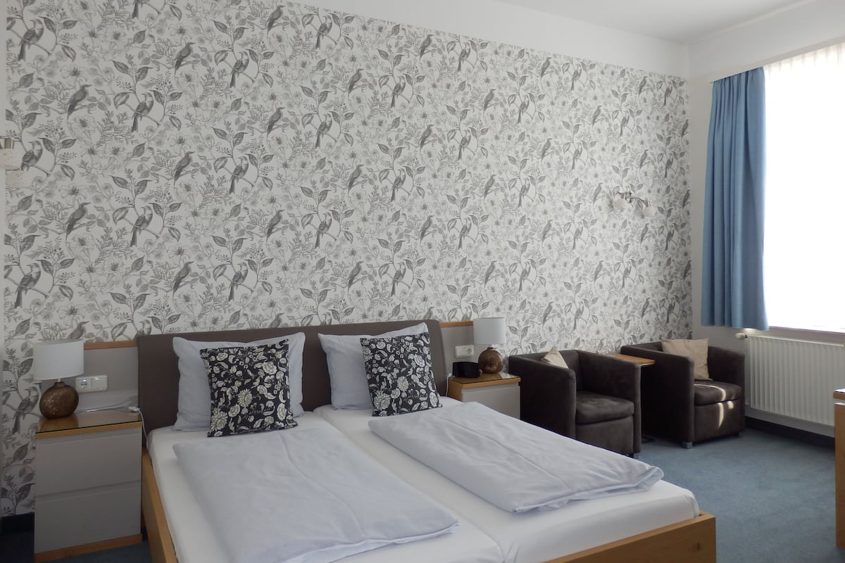 Ein Hotelzimmer mit einem großen Bett, zwei Nachttischen, einem Sessel und einem Fenster mit Vorhängen.