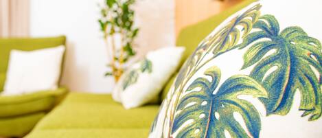 A room with a green sofa, a white pillow with a leaf pattern, and a plant in the background.