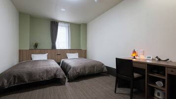 A hotel room with two single beds, a desk with a chair, a lamp, a clock, and a shelf with kitchenware.