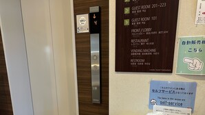 En hotellobby med en selvbetjent bagagevogn, et digitalt elevatordisplay og et skilt, der viser vejen til gæsteværelser og faciliteter.