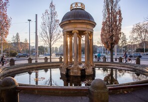 Una fuente clásica con una cúpula central y columnas, rodeada de una zona pavimentada con árboles y una calle al fondo.