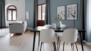 Un salón moderno con un sofá gris, una mesa de comedor con sillas blancas y un amplio ventanal con cortinas.