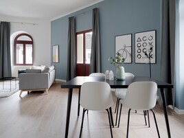 A modern living room with a grey sofa, a dining table with white chairs, and a large window with curtains.