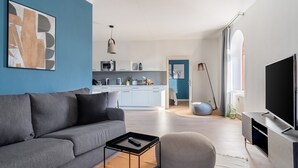 Una sala de estar moderna con un sofá gris, un pouf redondo, una mesa de centro y un televisor de pantalla plana. Se ve una cocina al fondo con una pared de acento azul.
