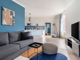 A modern living room with a grey sofa, a round ottoman, a coffee table, and a flat-screen TV. A kitchen is visible in the background with a blue accent wall.