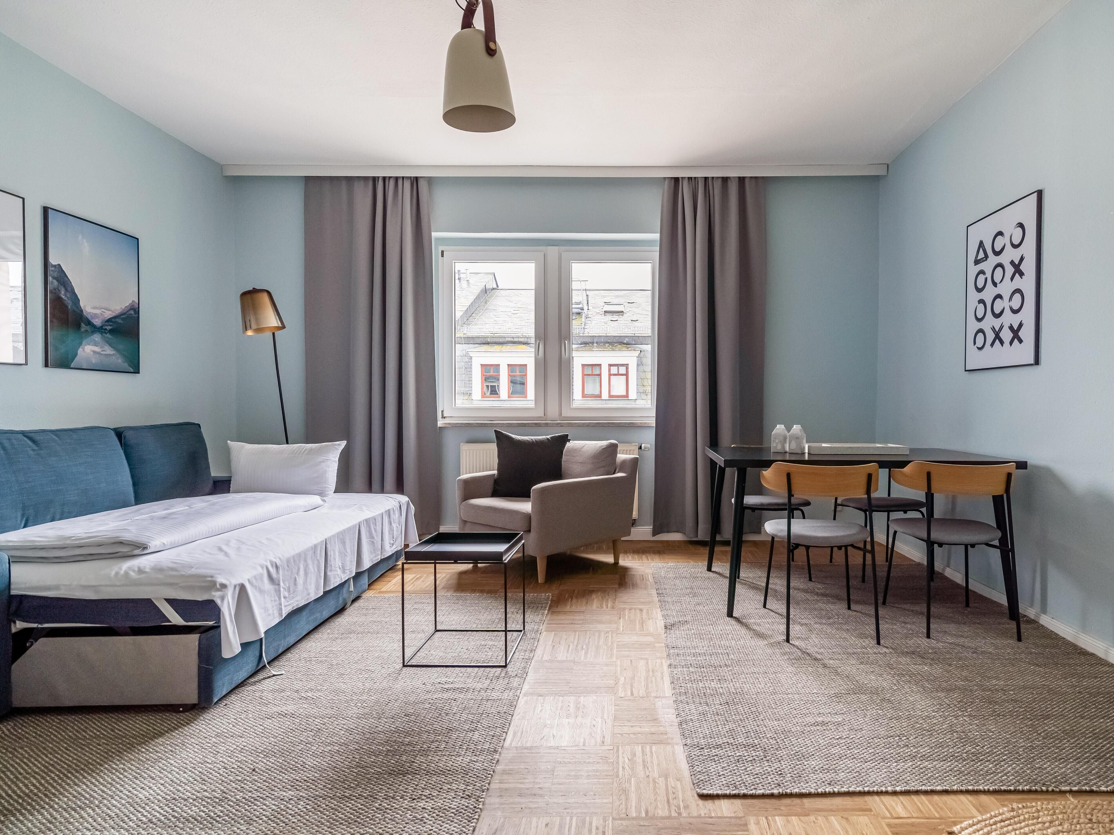 Ein modernes Schlafzimmer mit Bett, Schreibtisch und Stuhl. Ein Fenster mit Vorhängen, eine Lampe und ein Bild sind zu sehen.