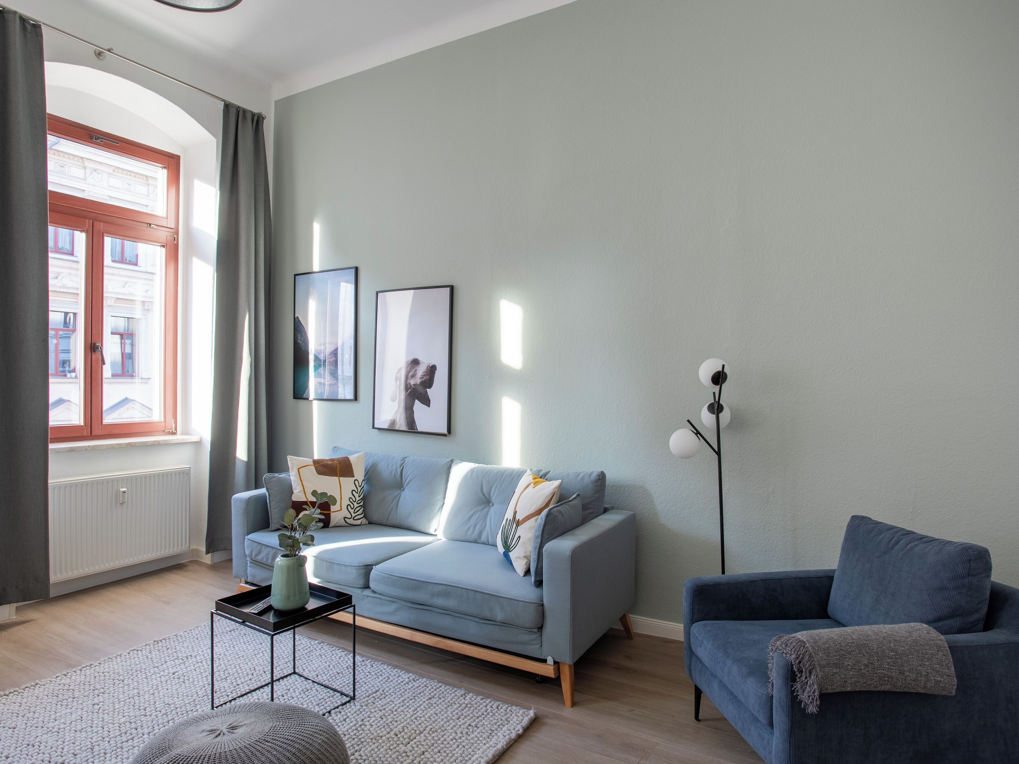 A modern living room with a light blue sofa, a dark blue armchair, a floor lamp, and two framed pictures on the wall.