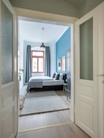 A bedroom with a bed, a window with curtains, a hanging light, and a door leading to another room.