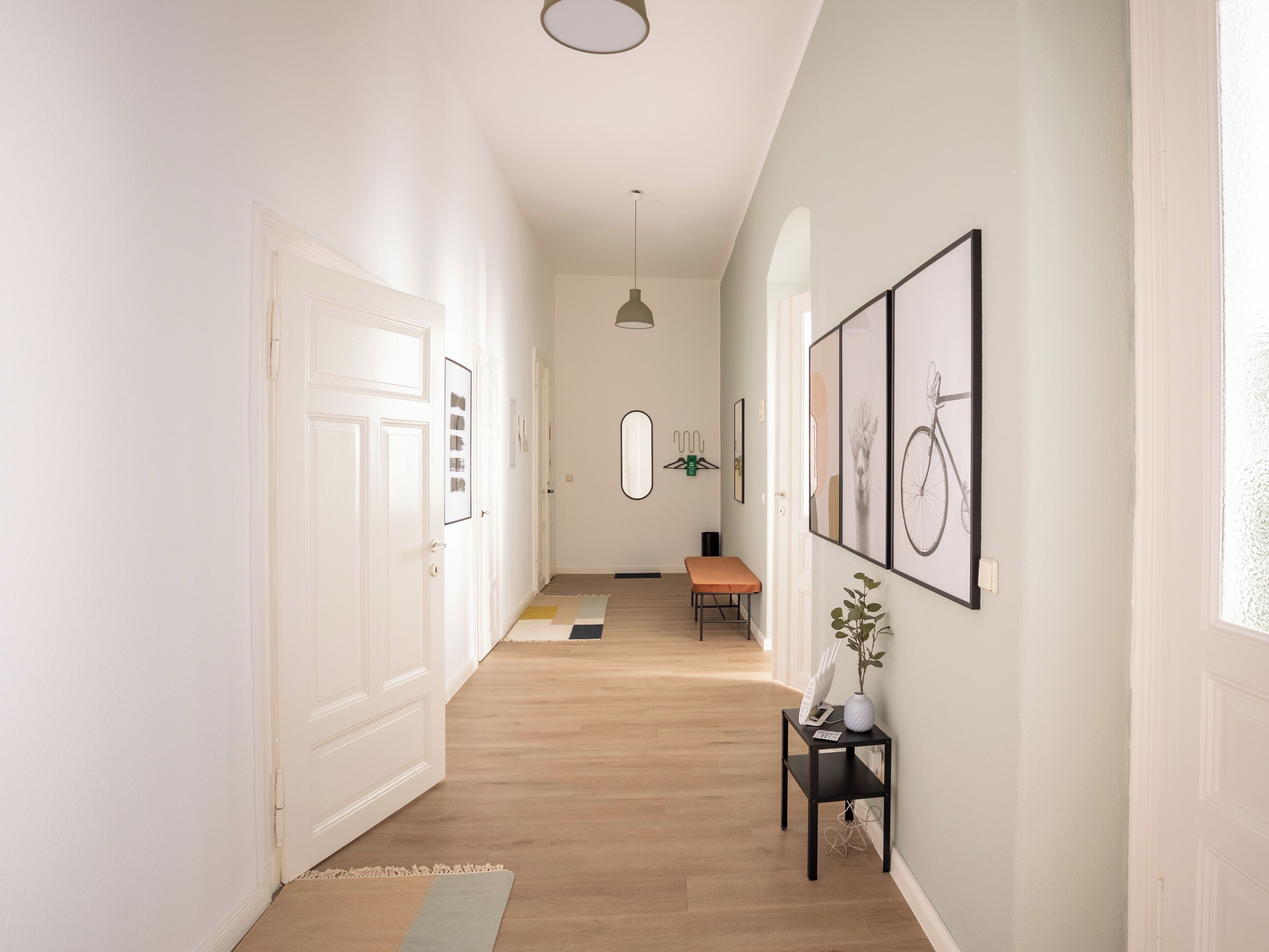 A hallway with a wooden floor, white walls, and a bench. There are framed pictures on the wall and a hanging light fixture.