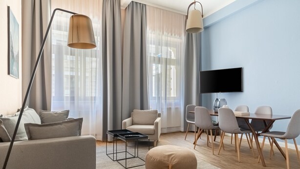 Una sala de estar moderna con un sofá, una mesa de centro, una mesa de comedor con sillas, un televisor montado en la pared y ventanas grandes con cortinas.