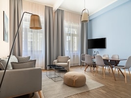 A modern living room with a sofa, a coffee table, a dining table with chairs, a TV mounted on the wall, and large windows with curtains.