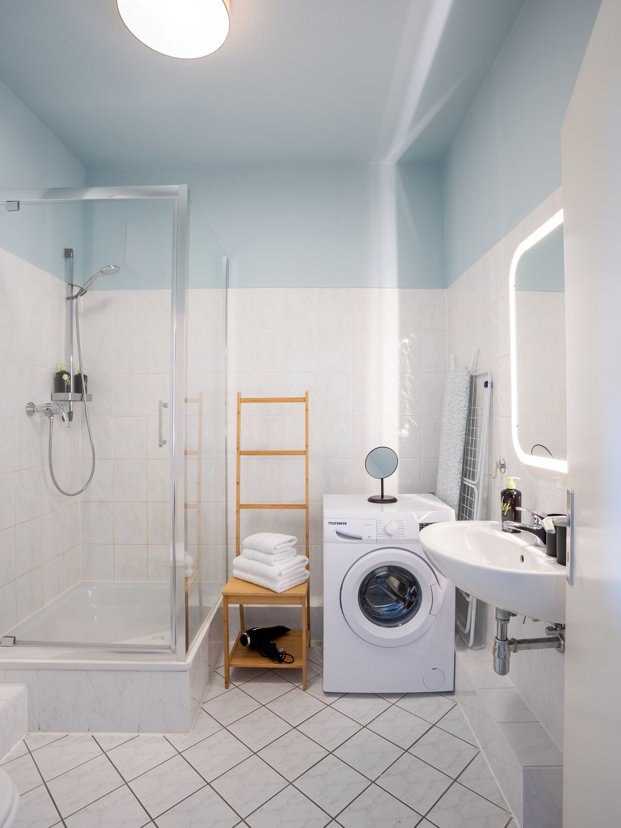 A modern bathroom with a glass shower enclosure, a wooden chair, a washing machine, and a sink with a mirror.