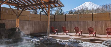 Uma área de águas termais com uma pérgola de madeira, cadeiras vermelhas e vista para uma montanha nevada.