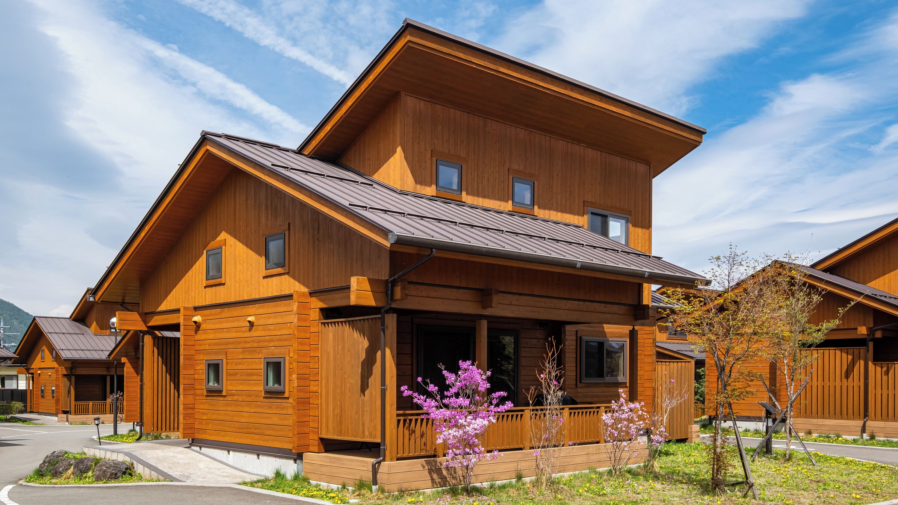 A two-story wooden house with a porch, a small garden with flowers, and a clear sky.