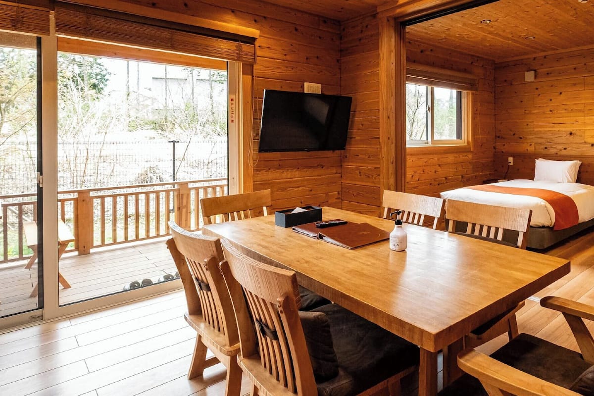 A wooden cabin with a dining table, two beds, a TV, and a balcony.
