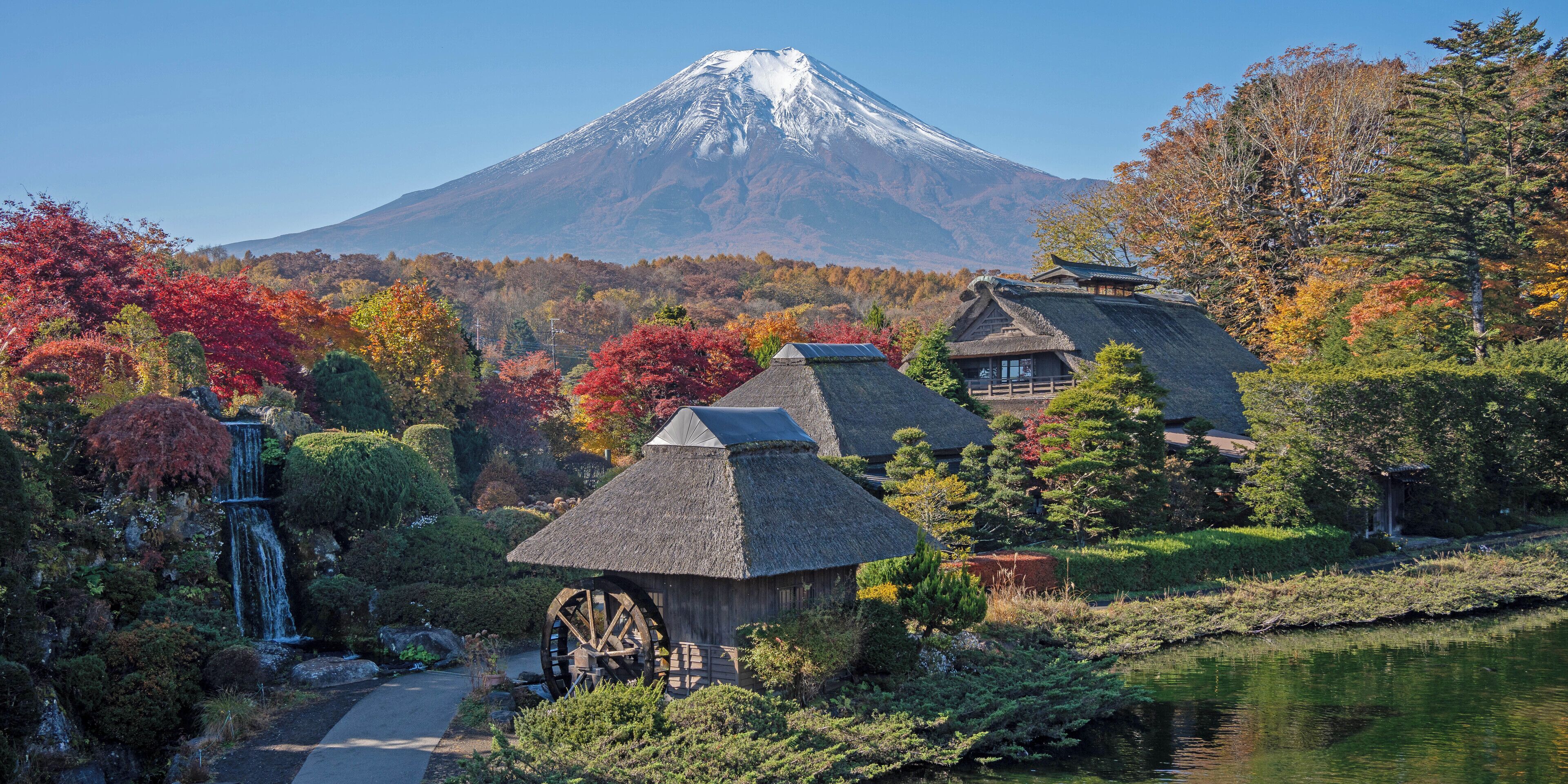 A traditional Japanese ryokan with thatched roofs, a water wheel, and a view of Mount Fuji in the background.