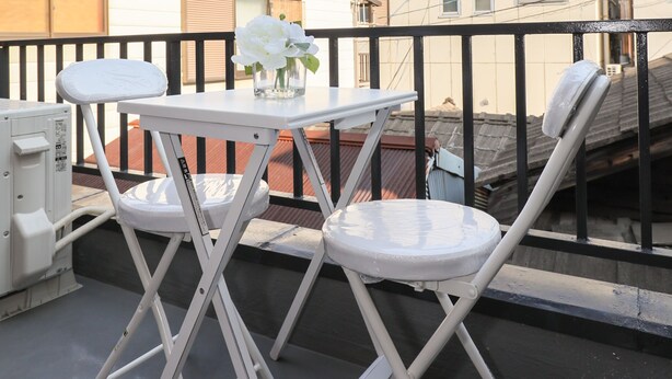 Un balcón con una mesa blanca y dos sillas blancas, un jarrón con flores blancas y vistas a edificios al fondo.