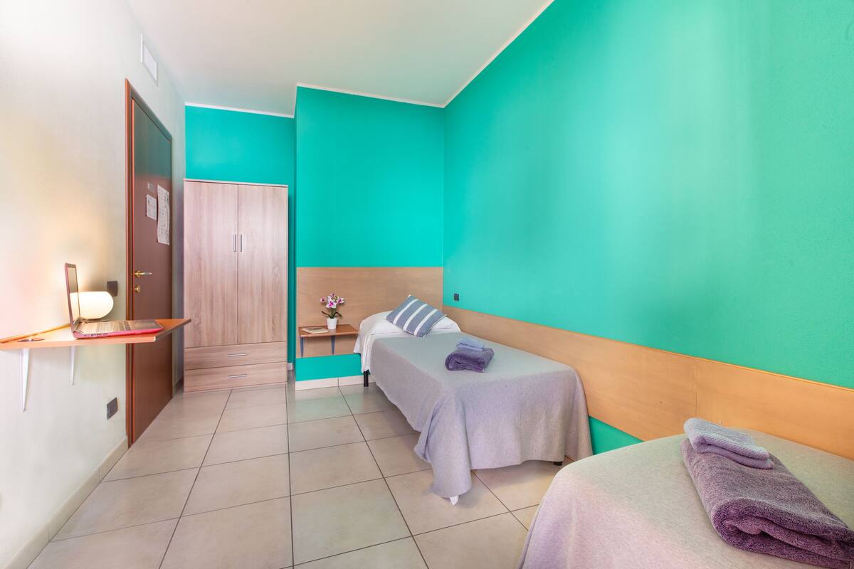 Habitación de hotel con dos camas, un armario de madera, un escritorio con una computadora portátil y una mesita con un jarrón.