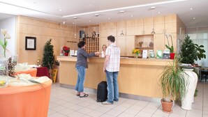 A reception area with a wooden counter, a receptionist, and a guest standing. There are potted plants and a table with a red tablecloth. - Behringers Freizeit- & Tagungshotel (Goessweinstein)