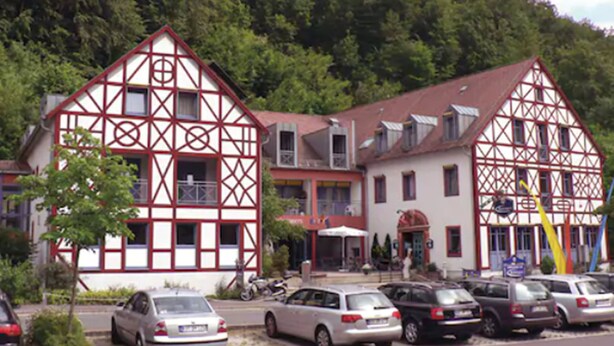 A traditional half-timbered building with a red and white facade, a parking lot with several cars, and a forested hillside in the background. - Behringers Freizeit- & Tagungshotel (Goessweinstein)