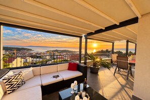 Un balcon aménagé avec un canapé, des chaises et une table, prêt pour un repas, offrant une vue sur le paysage urbain au coucher du soleil.