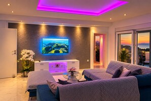 Een moderne woonkamer met een flatscreen televisie aan een gestructureerde muur, een zithoek, een salontafel en uitzicht op de omgeving via een glazen deur.