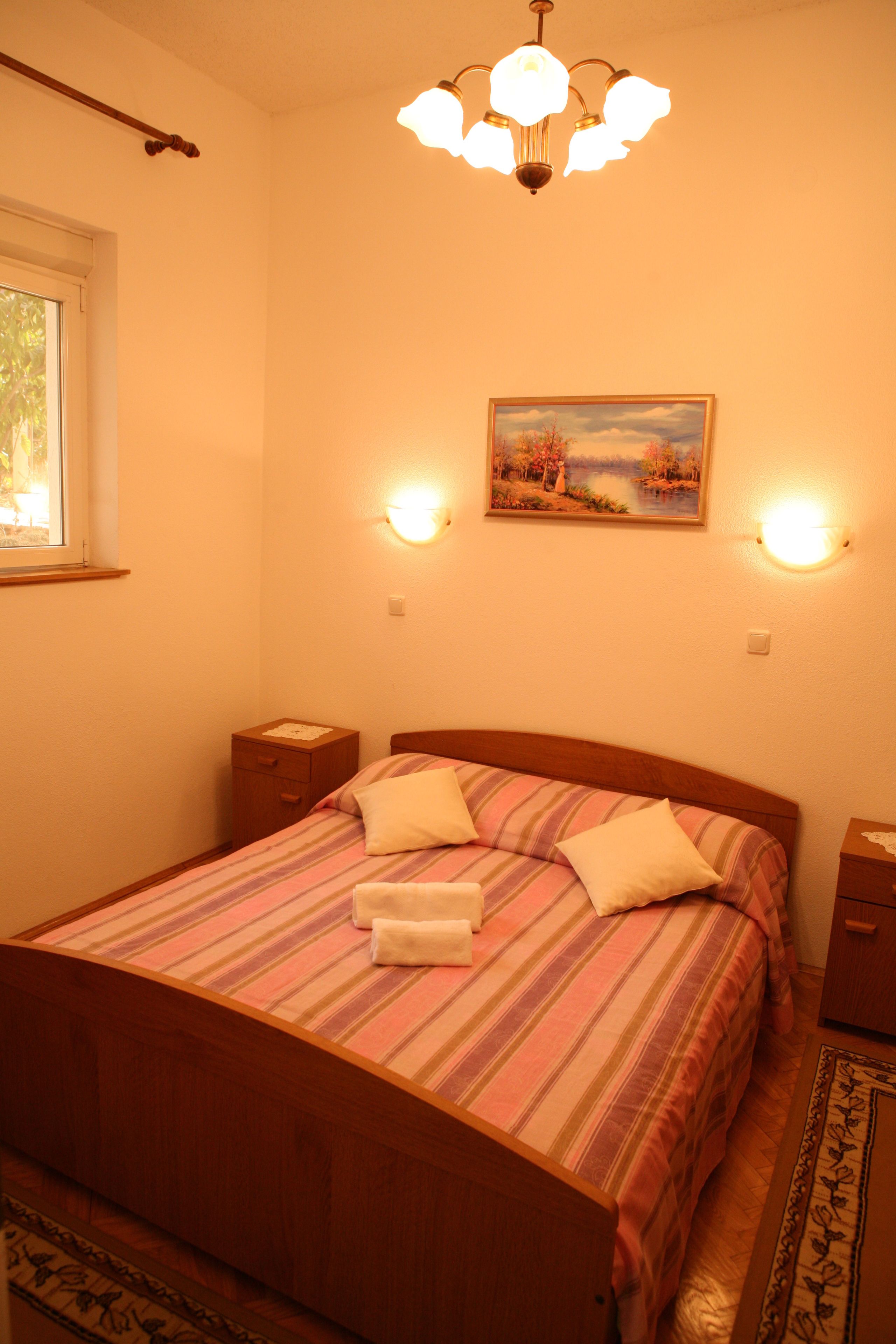 A bedroom with a wooden bed, a nightstand, a bedside table, a painting, and a chandelier.