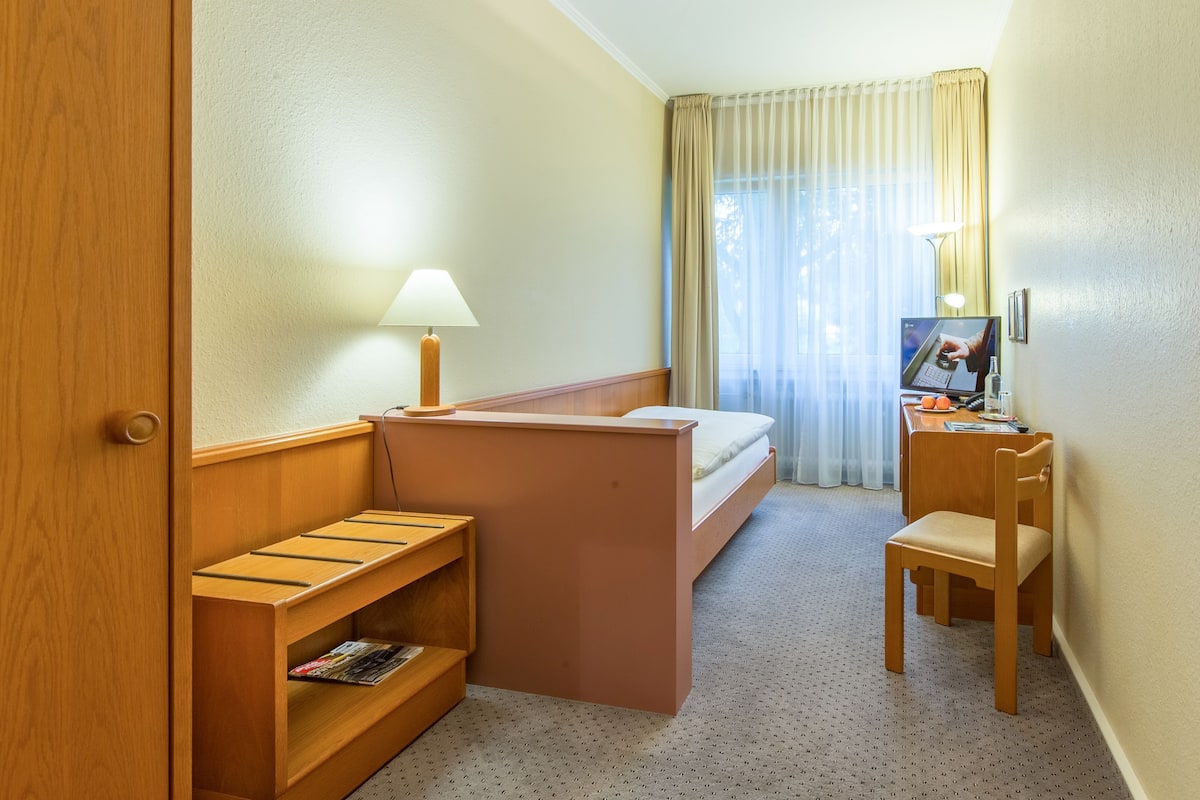 Une chambre d’hôtel avec un lit, un bureau avec un ordinateur, une chaise, une petite étagère et une fenêtre avec des rideaux.
