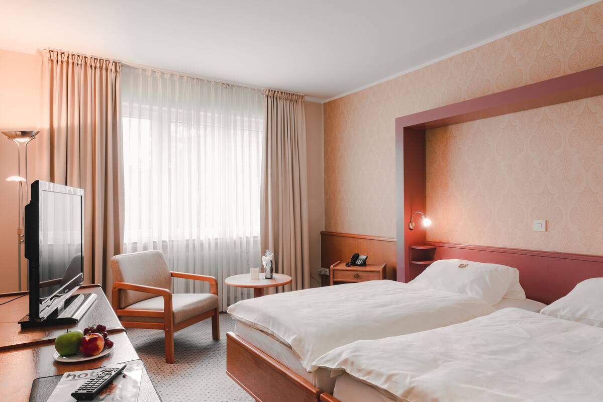 Une chambre d’hôtel avec deux lits, un bureau avec un ordinateur, une chaise, une télévision et une petite table sur laquelle se trouve une assiette de fruits.