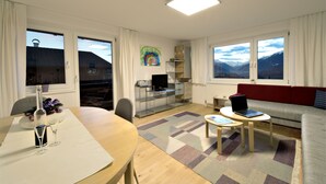 Una sala de estar moderna con comedor, un sofá rojo, un televisor y vistas a la montaña a través de grandes ventanales.