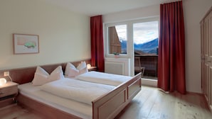 Una cama doble con cabecera de madera, una mesita de noche con lámpara, una ventana con cortinas rojas y vistas a las montañas.