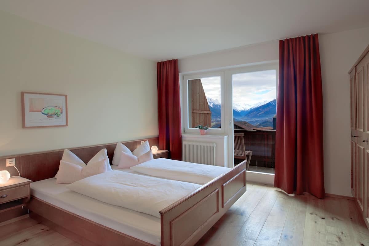 Una cama doble con cabecera de madera, una mesita de noche con lámpara, una ventana con cortinas rojas y vistas a las montañas.