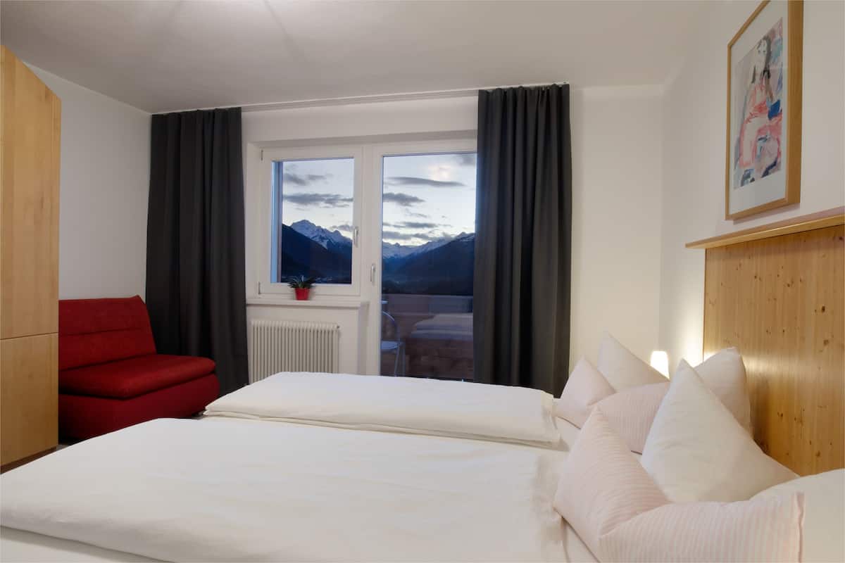 Una habitación de hotel con una cama, una silla roja, una ventana y vista a las montañas, y un cuadro en la pared.
