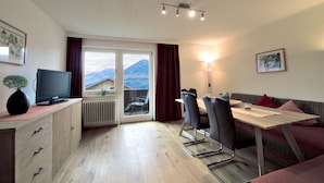 Una sala de estar moderna con televisión, mesa de comedor y sofá. Cuenta con un ventanal con vistas a la montaña.