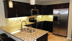 A modern kitchen with dark cabinetry, a stainless steel refrigerator, granite countertops, and a hanging light fixture.