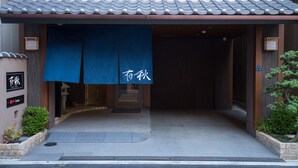 A building entrance with a blue awning, a tiled roof, and a small garden area with plants. - Hotel Yu-shu (Osaka)