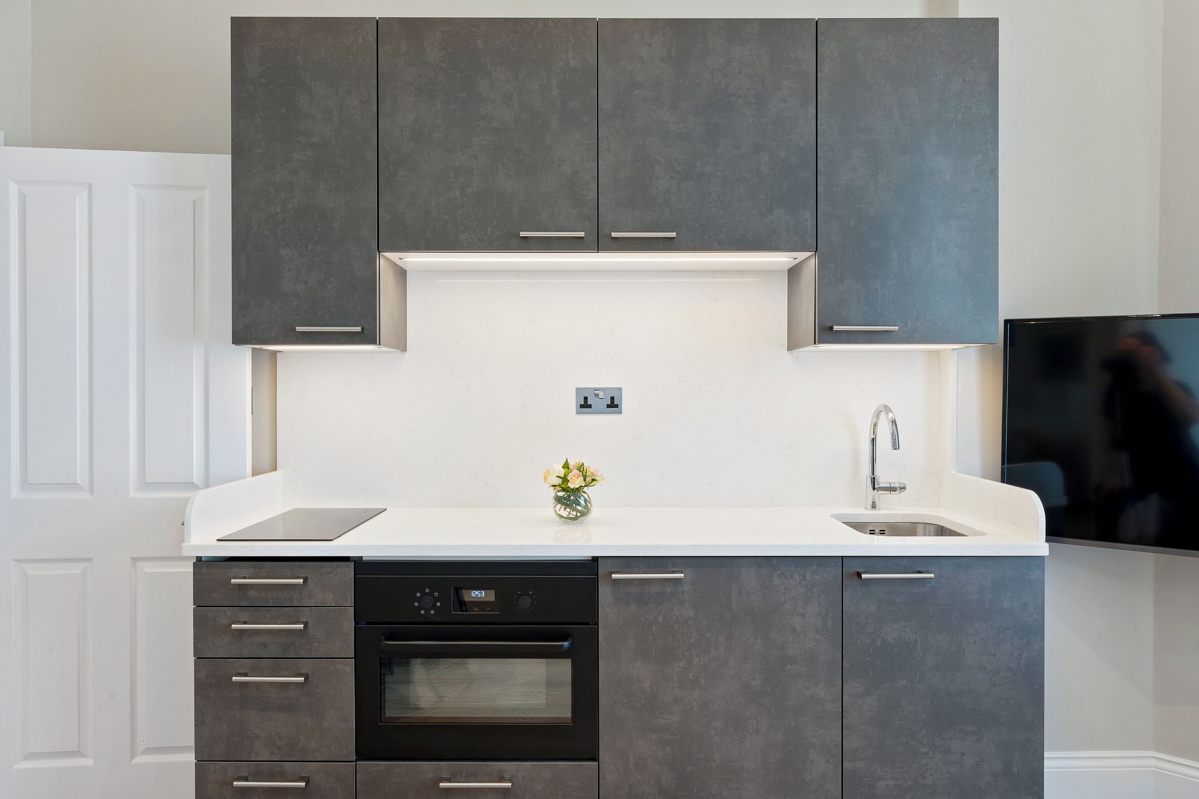A modern kitchen with dark cabinets, a white countertop, a built-in oven, a dishwasher, and a small vase with flowers.