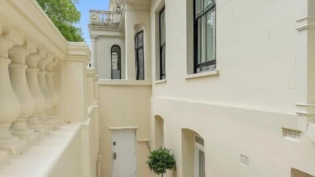 A narrow, tiled corridor with white walls and a balustrade on the left. There are two potted plants and a door at the end of the corridor. - Cleveland Residences Paddington (London)