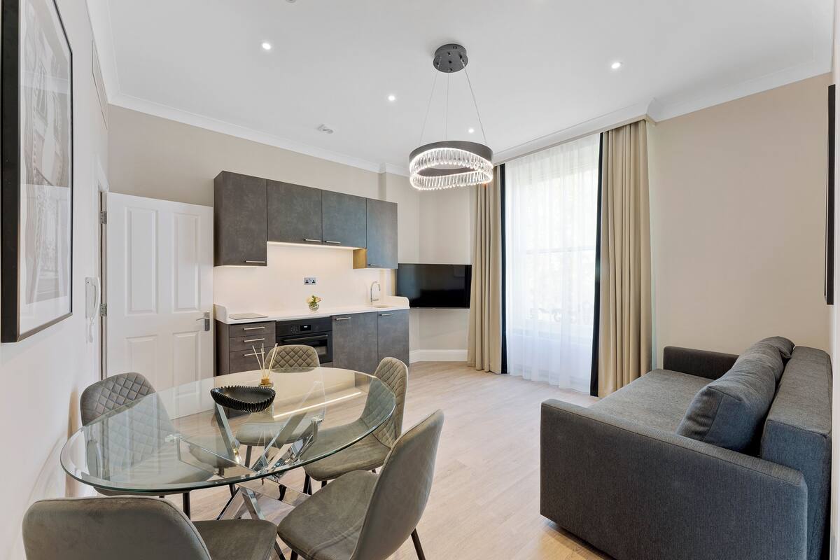 A modern living room with a glass dining table, grey sofa, and a kitchen area with dark cabinets and a flat-screen TV.