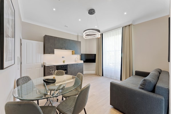 Un salón moderno con una mesa de comedor de cristal, sofá gris y una zona de cocina con armarios oscuros y un televisor de pantalla plana.