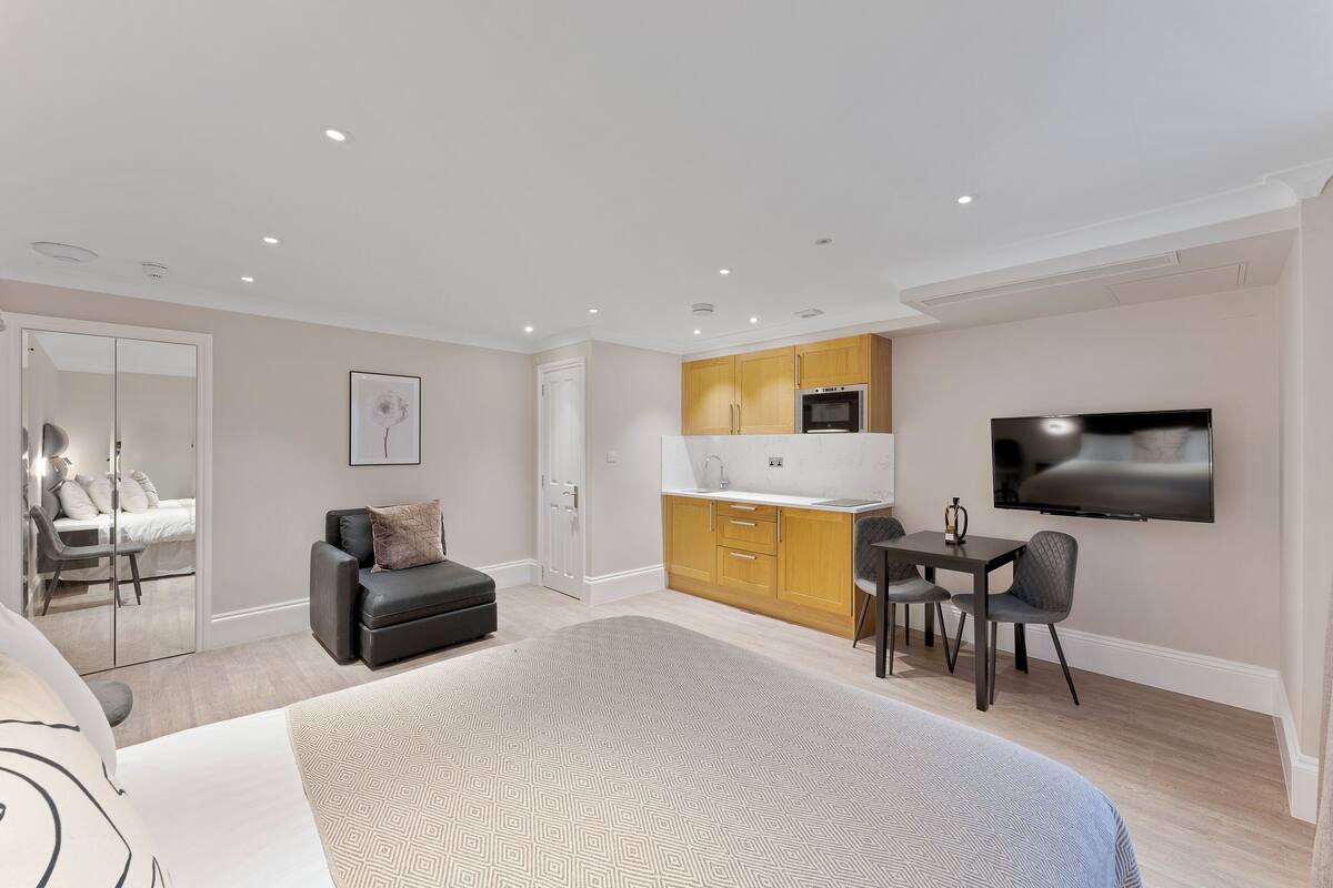 A modern hotel room with a large bed, a small desk, a flat-screen TV, a kitchenette, and a separate bathroom.