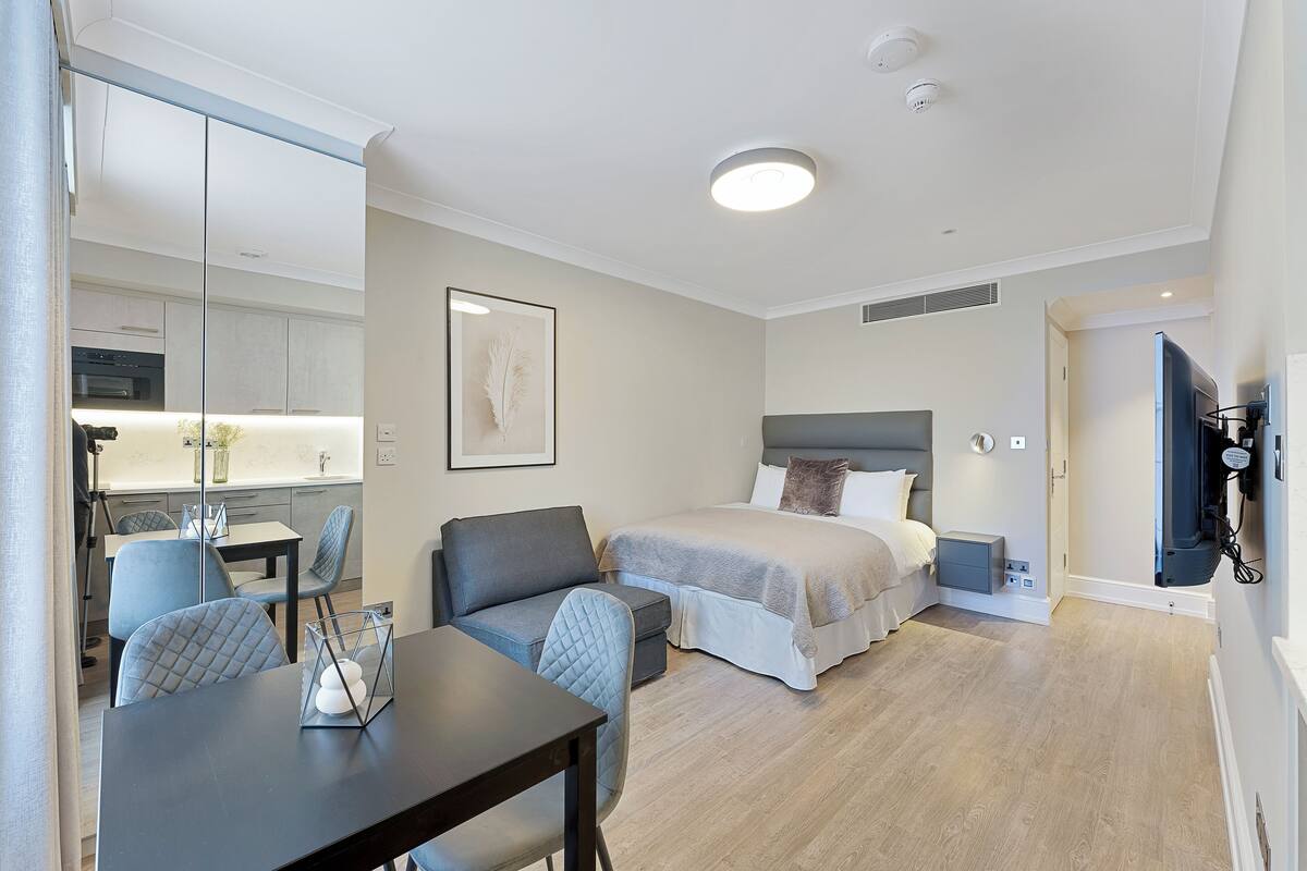 A modern hotel room with a bed, bedside table, TV, and a dining area with a table and chairs.