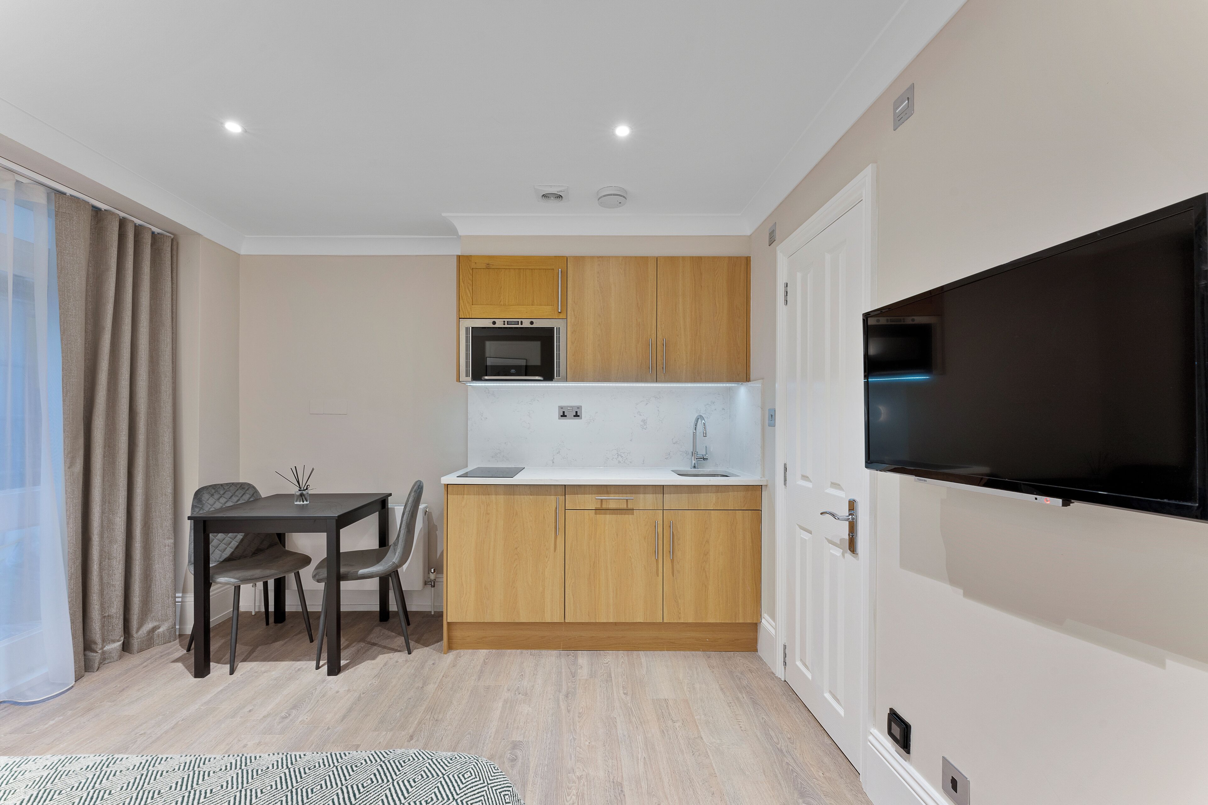 A compact kitchen with wooden cabinets, a microwave, a sink, and a large TV mounted on the wall.