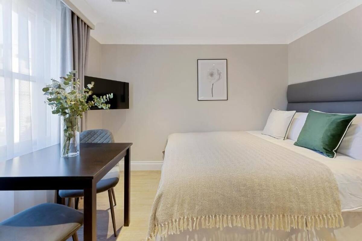 A modern hotel room with a large bed, a flat-screen TV mounted on the wall, a dining table with chairs, and a vase with flowers on the table.