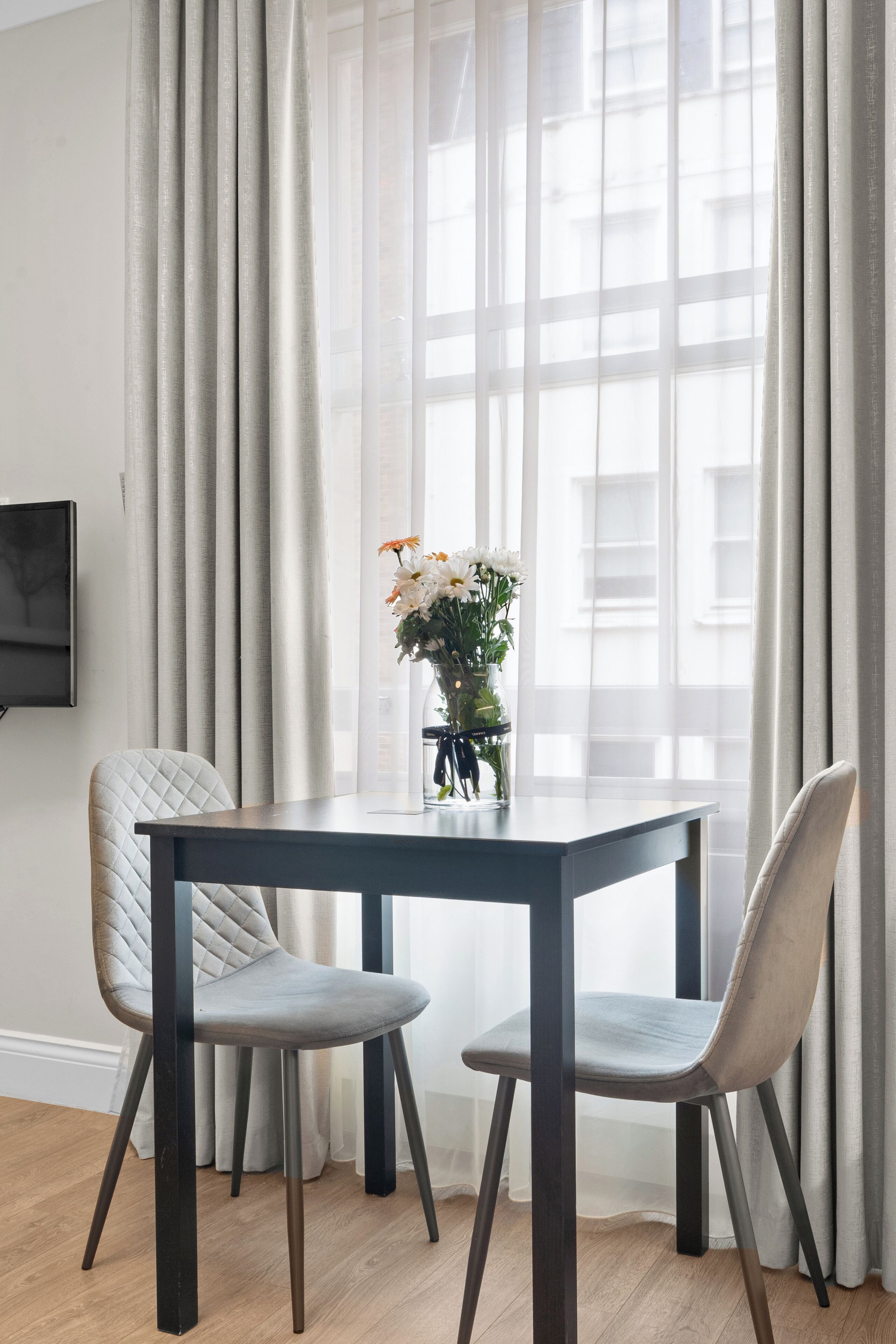 Een eethoek met een tafel en twee stoelen, een vaas met bloemen en een raam met gordijnen.