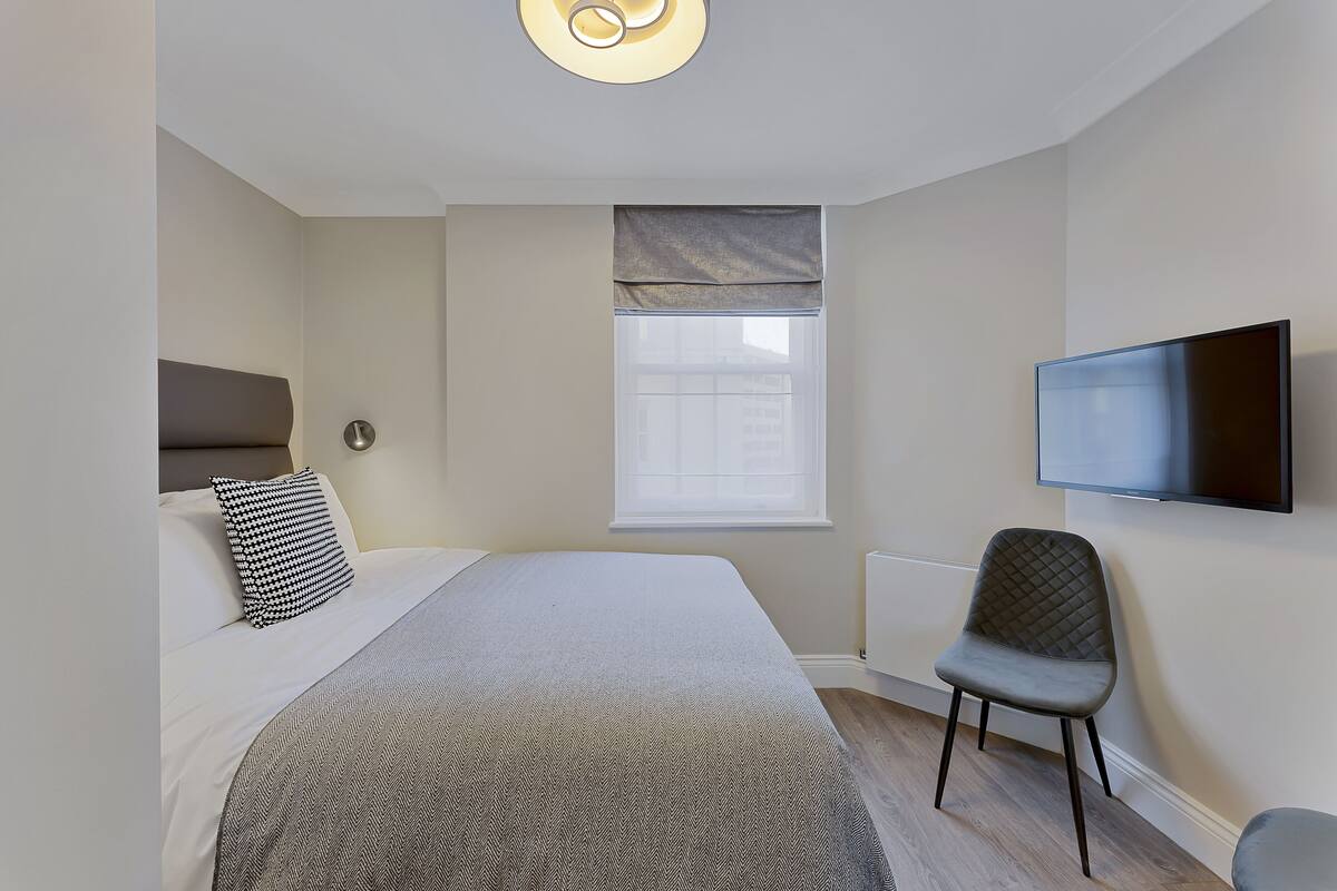 A bedroom with a bed, a chair, a television, and a window with blinds.